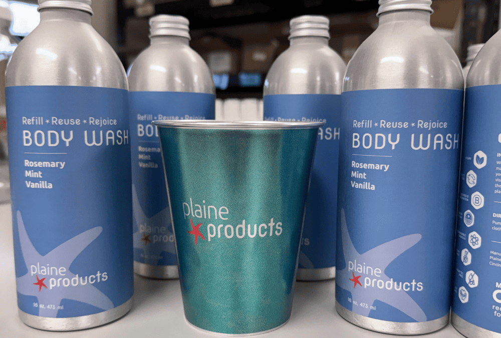 A customized LumiCup with the Plaine Products logo, pictured with bottles of Plaine Products Rosemary Mint Vanilla Body Wash.