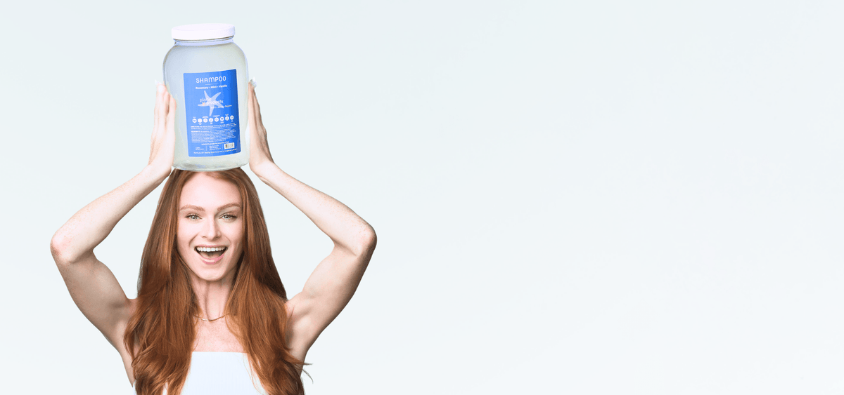A woman with long red hair holds a large gallon jar of Plaine Products Shampoo on her head against a light gray studio background.